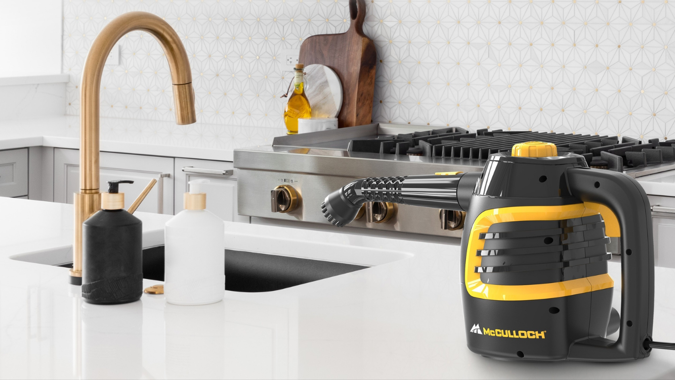 A McCulloch steam cleaner in black and yellow sits on a white kitchen counter next to a gold faucet, with a gas stove and kitchen backsplash in the background.